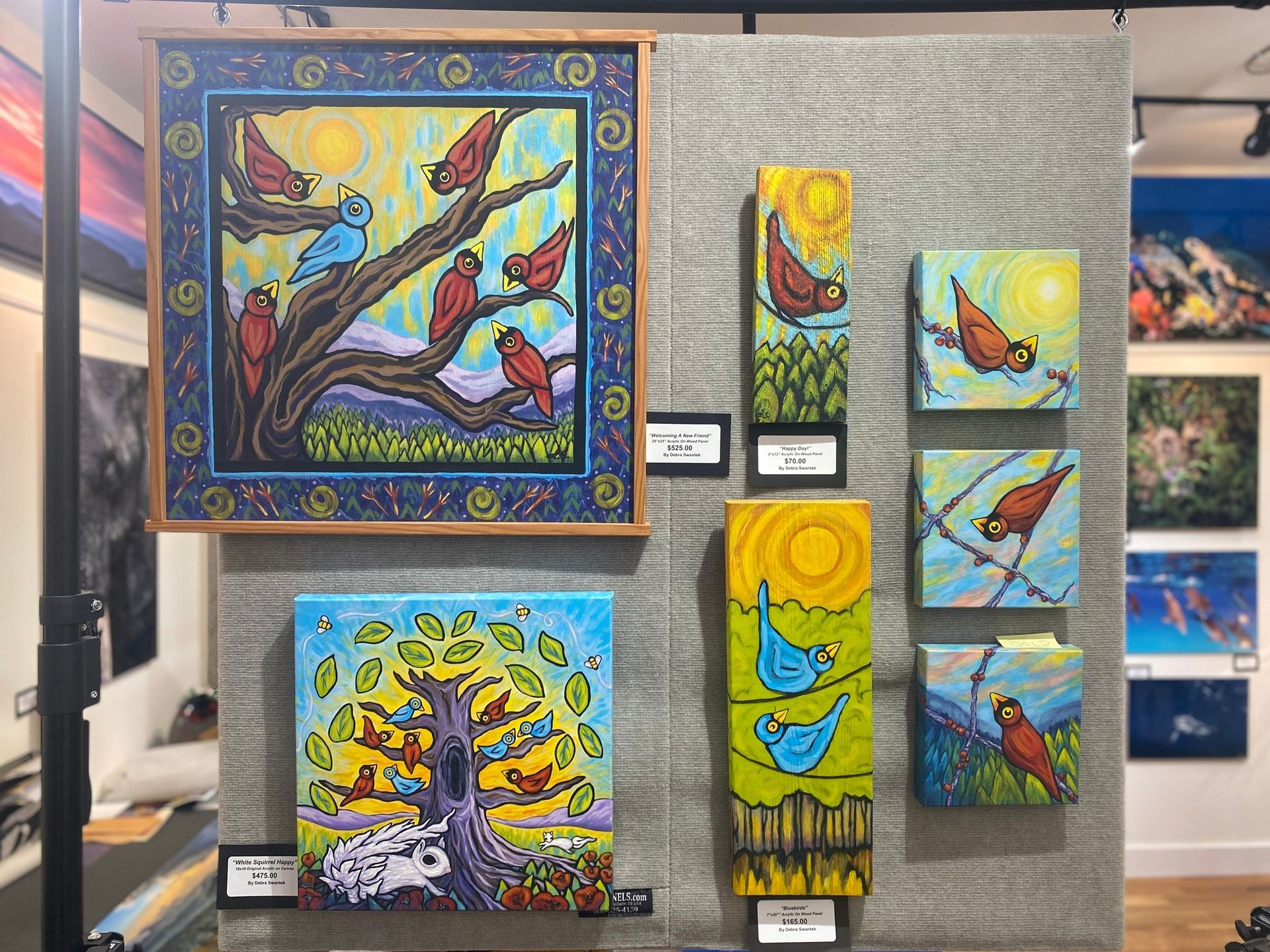 Colorful abstract paintings of birds displayed on a gallery wall with bright blues, yellows, and reds.