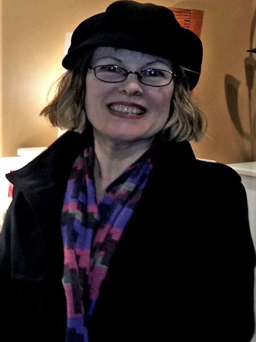 Smiling person in a black hat and coat with a colorful scarf, standing indoors.