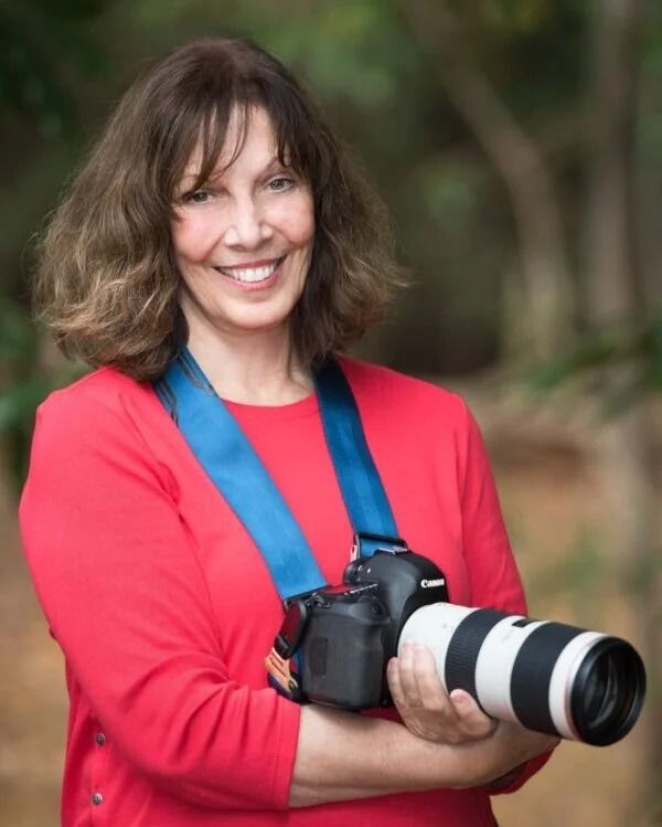 Smiling woman in red shirt holding a DSLR camera with a large white telephoto lens outdoors