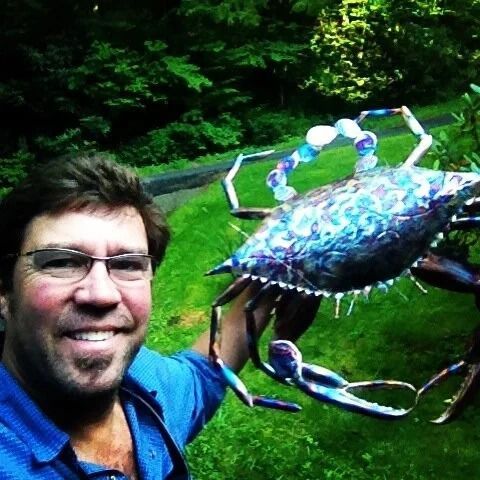 Man smiling on a trail, holding a large blue crab up close in a lush green forest