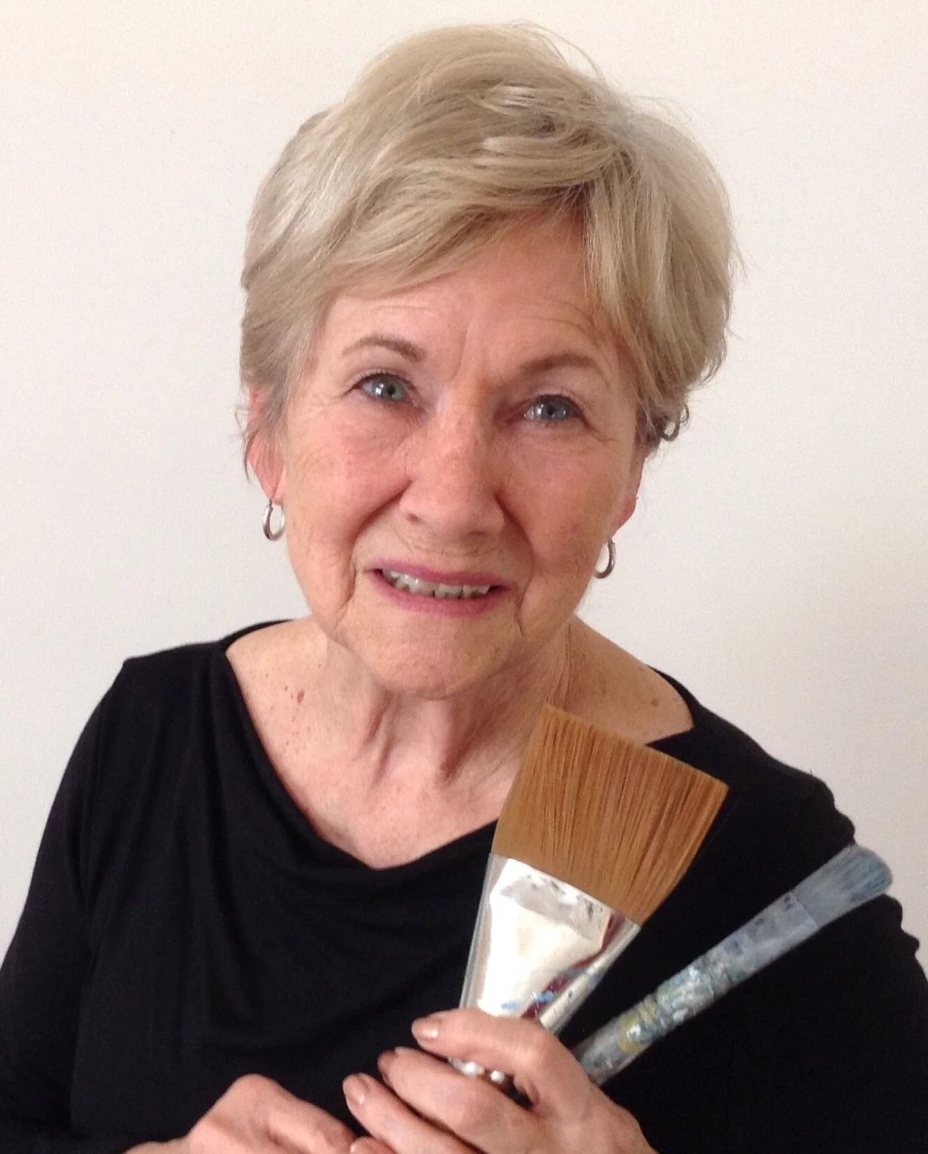 Smiling woman holding a makeup brush and a tube of product against a plain background