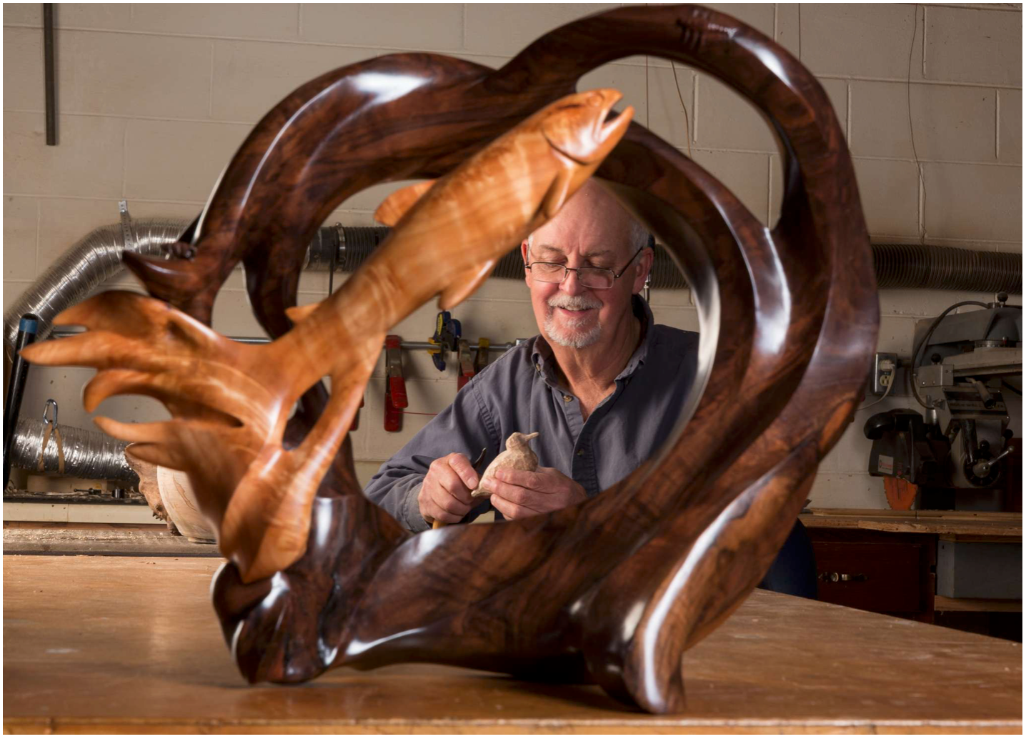 Wooden sculpture with intertwined branches on a table, with a man smiling behind it in a workshop.