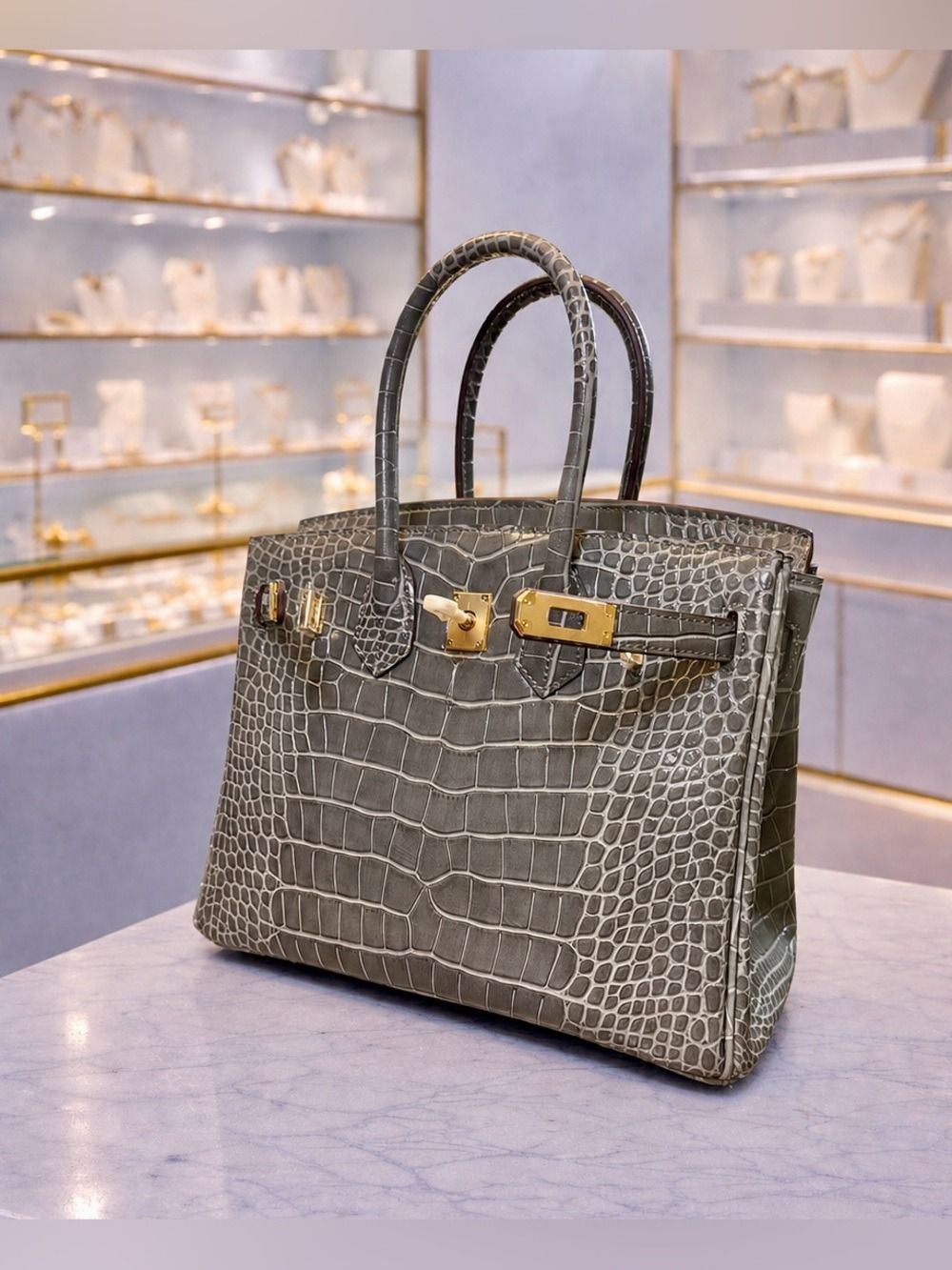 Gray crocodile-textured handbag with gold clasps on a marble table in a softly lit boutique display