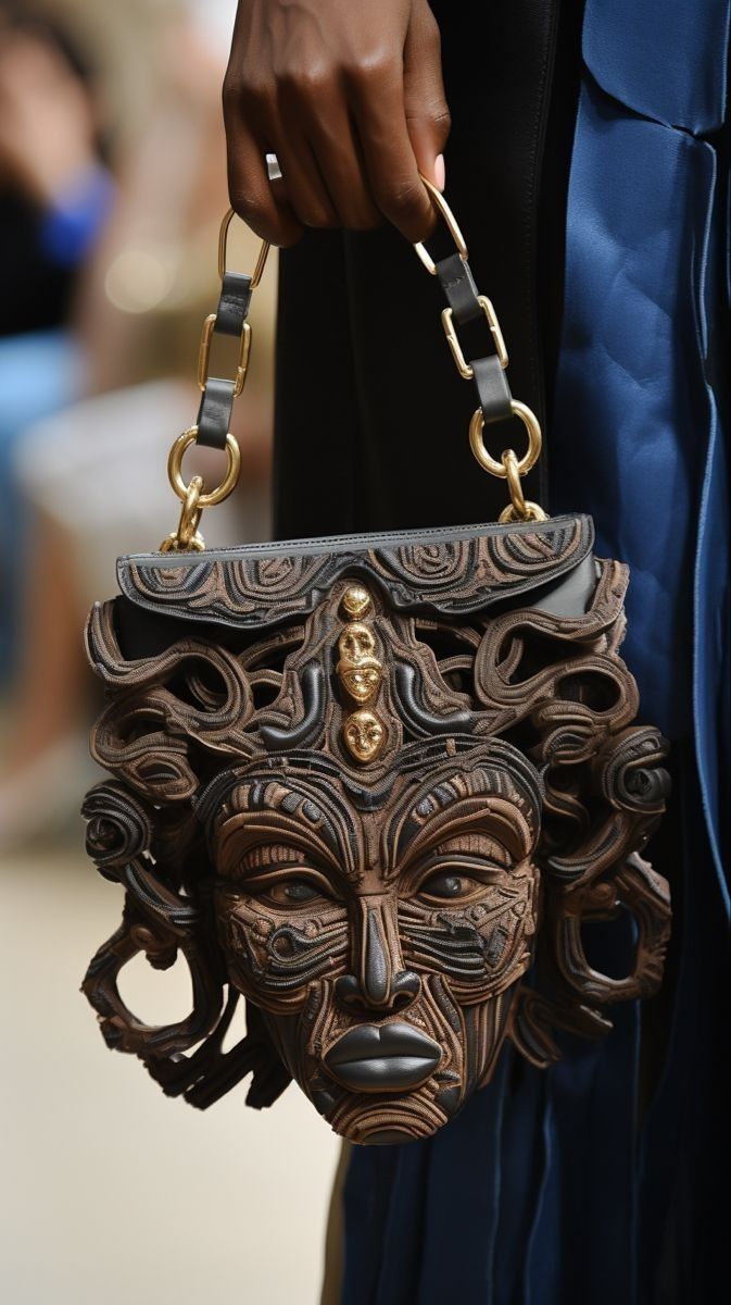 Hand holding a dark carved mask-shaped handbag with chain straps against a blue dress