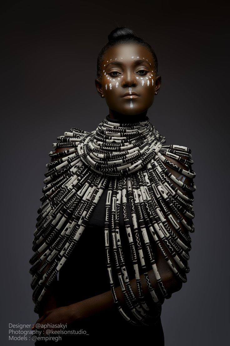 Person in dark, sculptural metal-like garment against a black background, facing forward.