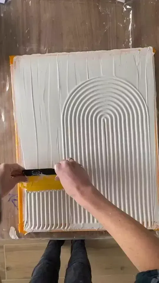 Hands spreading white adhesive on a board with a notched trowel, creating curved ridges.