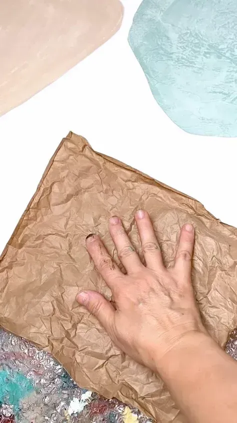 Hand smoothing a crumpled brown paper sheet on a white surface with pastel paint patches.
