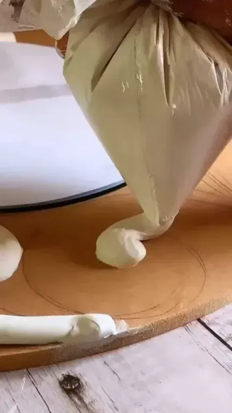 White icing being piped onto a wooden board next to a partially frosted cake.