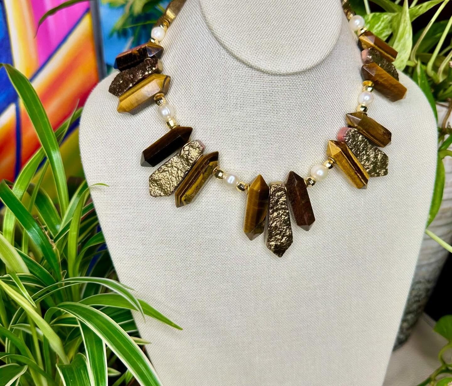 White bust display with a gold and brown statement necklace, beside green leaves and a colorful background