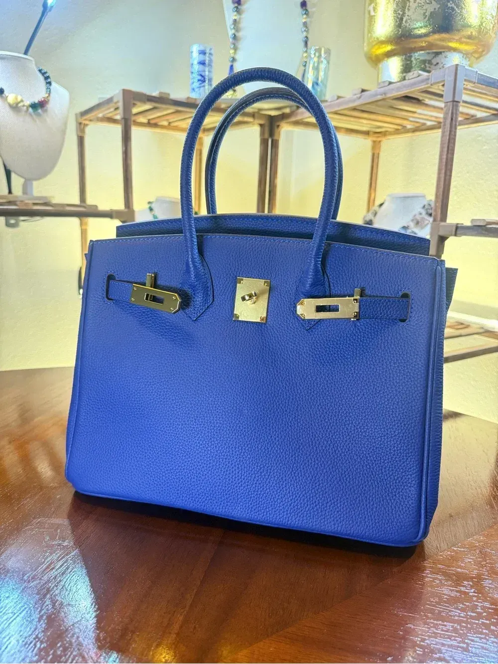 Blue handbag with top handles and gold hardware on a wooden table, in a bright room