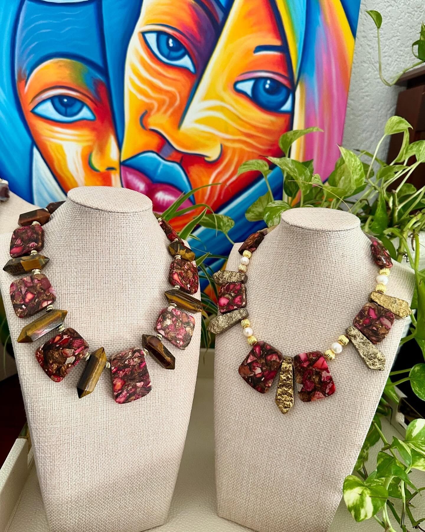 Two beaded necklaces on display busts, with a colorful mural and green plants behind them.