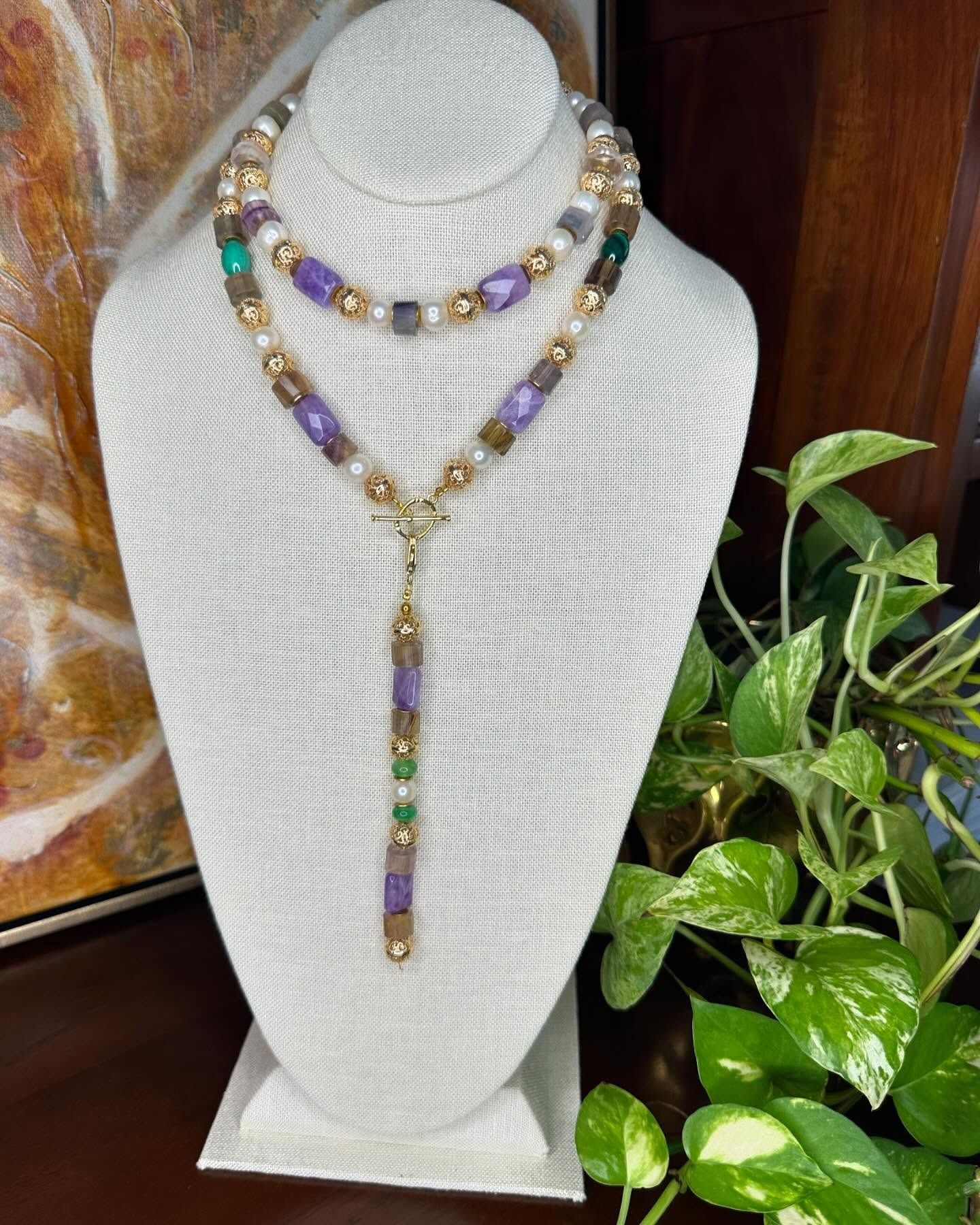 Purple and gold beaded necklace and lariat on a white display bust beside a green plant