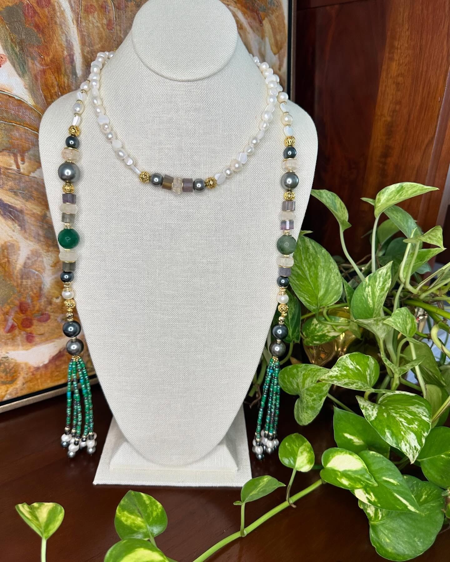 Beaded necklace on a white bust display with green and black accents, beside a leafy plant.