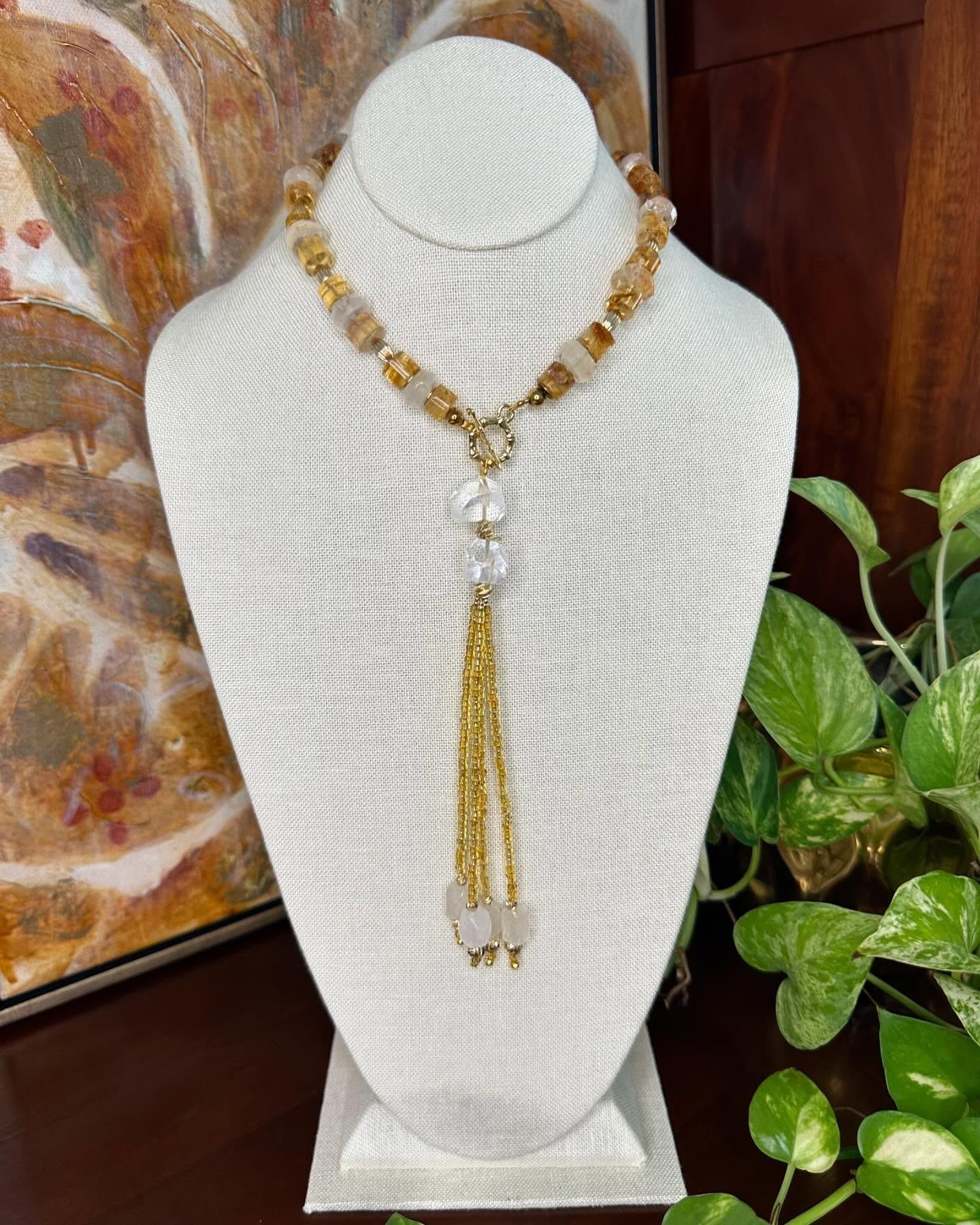 White necklace bust displaying a gold beaded necklace with a long tassel pendant, beside a green plant