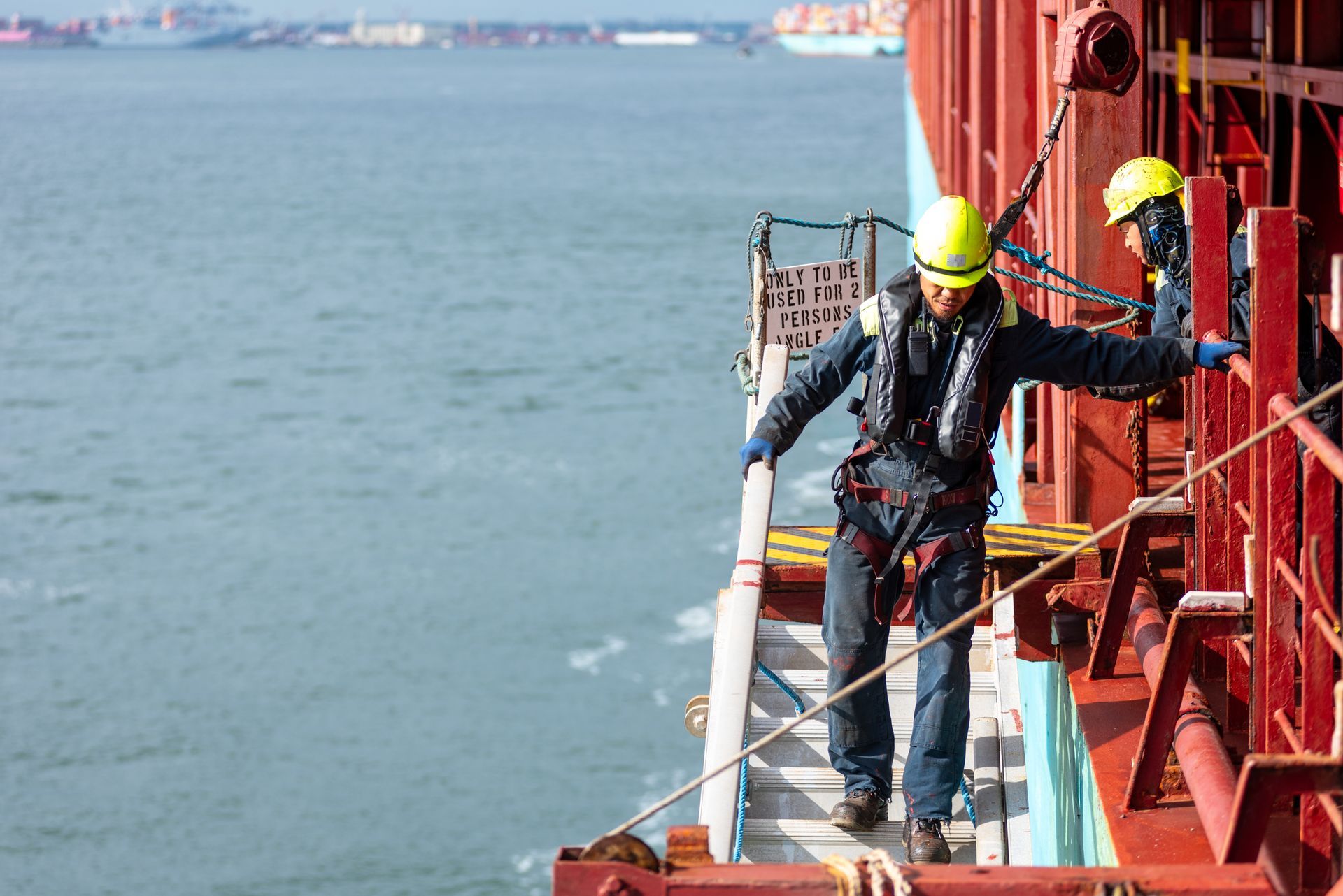 Two people in safety gear on a ship, near the ocean. One walks down steps.