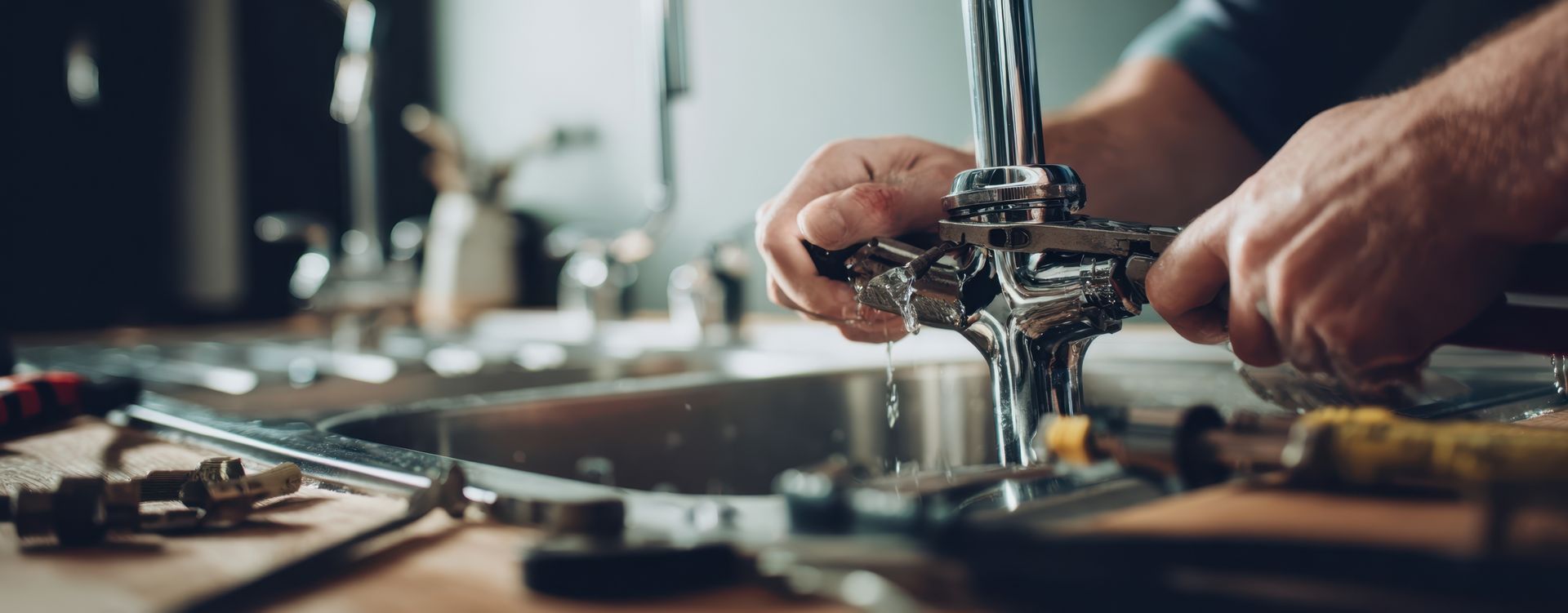 A person's hands fixing a leaky faucet. Tools are scattered nearby on a wooden surface.