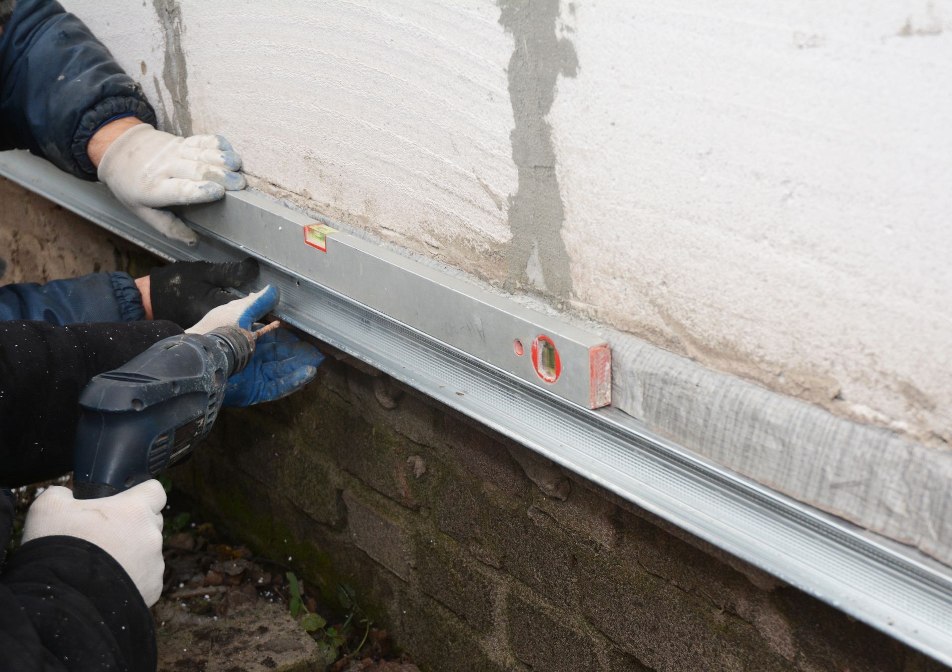 Two people installing a metal track on a concrete wall using a level and power drill outdoors.