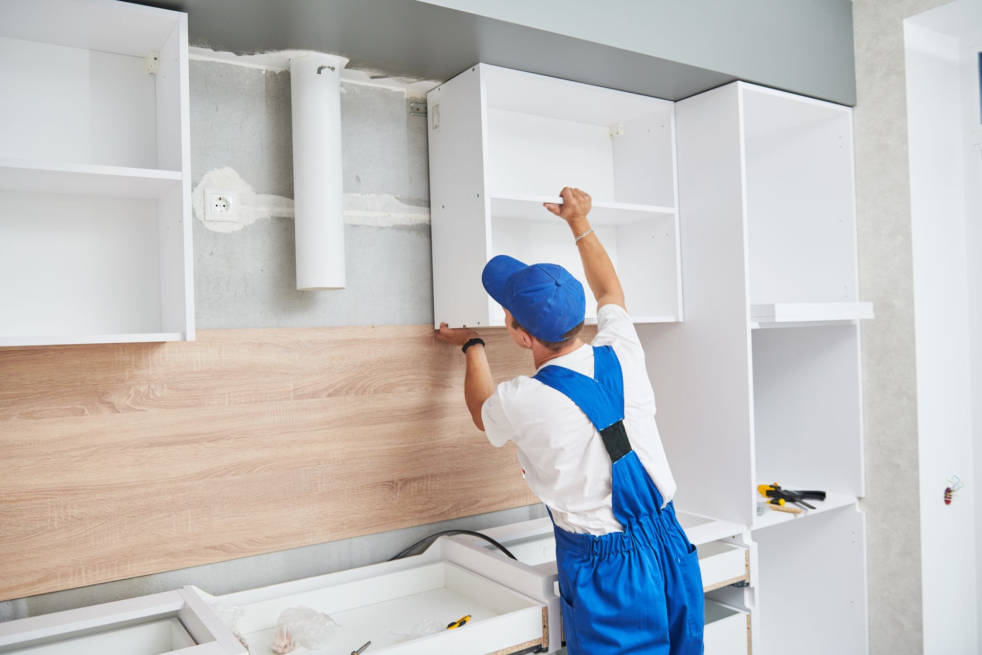 Person in blue overalls installing white kitchen cabinet, indoors.