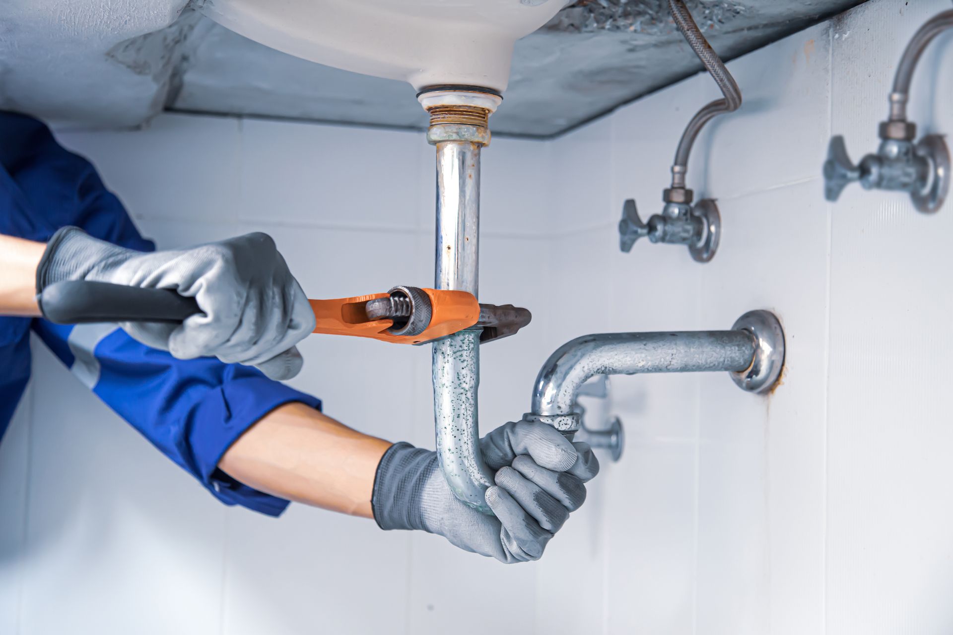 Plumber with wrench fixing sink pipes under a white sink in a tiled room.