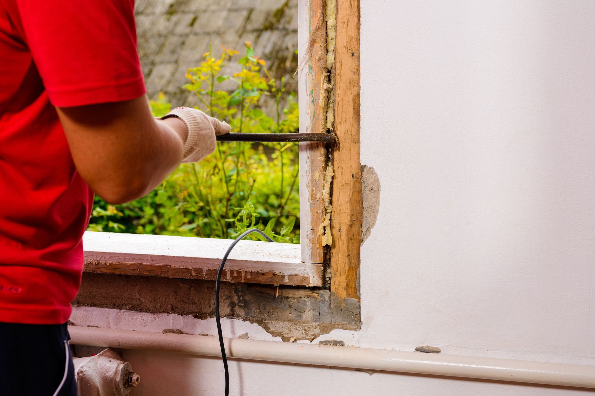 Person removing a window frame with a tool. The person wears a red shirt, working near a green backdrop.