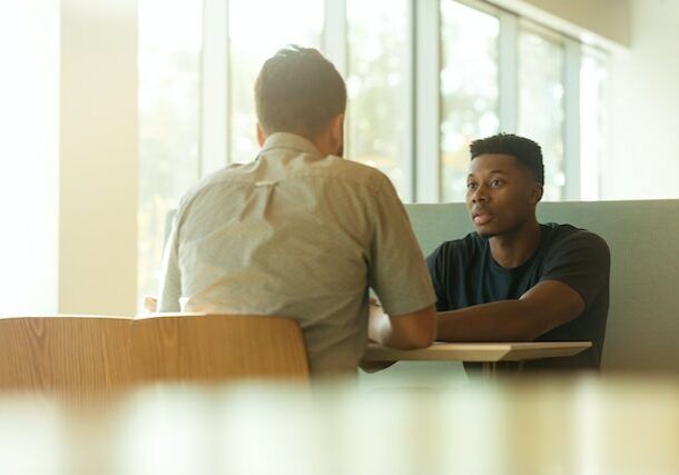 Two men in a booth; one faces the viewer, talking, while the other's back is turned. They sit at a table near a window.