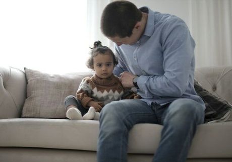 Man in blue shirt and jeans interacting with a young child on a couch indoors. The child wears a sweater.