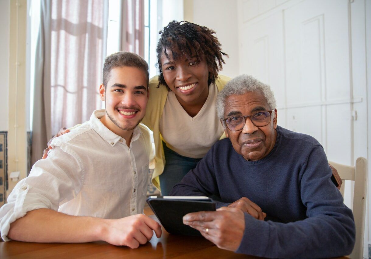 Three people, a young man, a young Black woman, and an elderly Black man, smile while gathered around a tablet at a table.