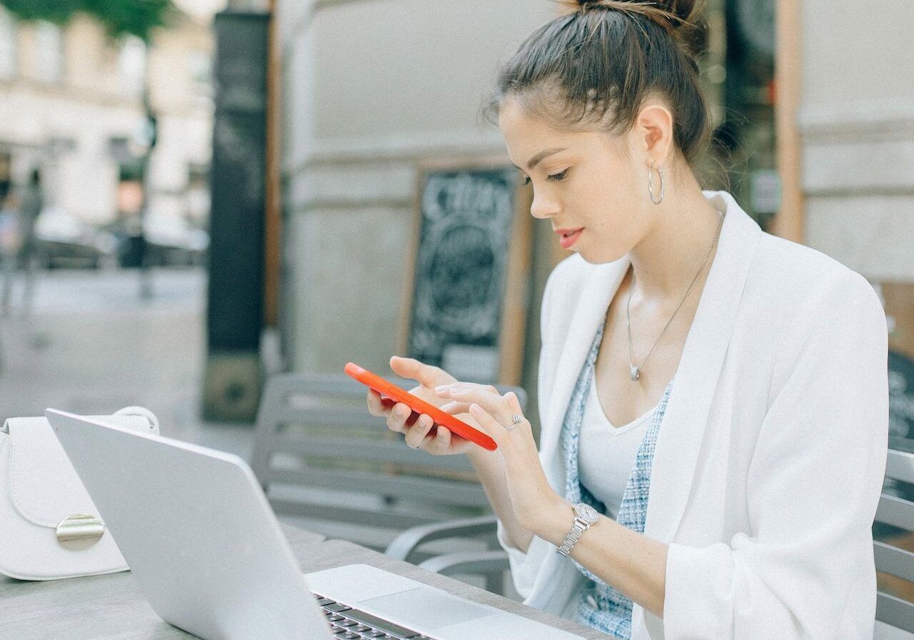 Woman in white blazer uses a red phone at an outdoor table with a laptop and purse.