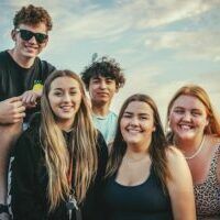 Group of young people smiling, posing outdoors under a blue sky; includes two girls in the front and others in the background.