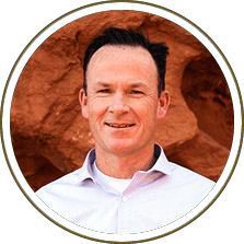 Portrait of a smiling man in a white shirt, framed by a circular border against a red rock background