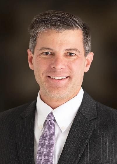 Studio headshot of a man in a dark suit and purple tie, smiling against a blurred dark background
