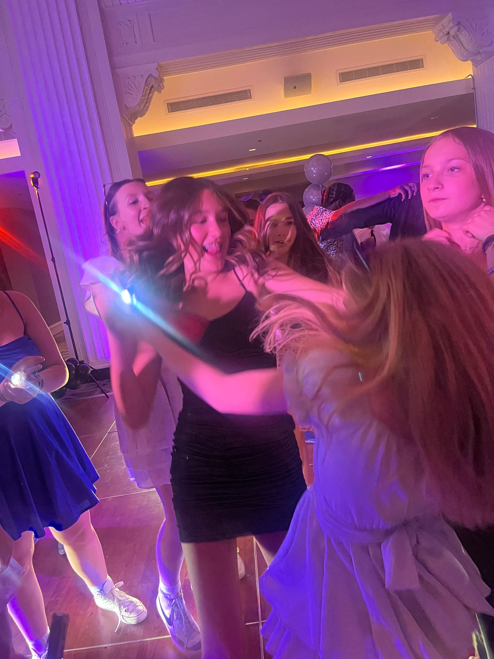 Teenagers dancing at a party with purple lighting. Several girls are wearing dresses and having fun.
