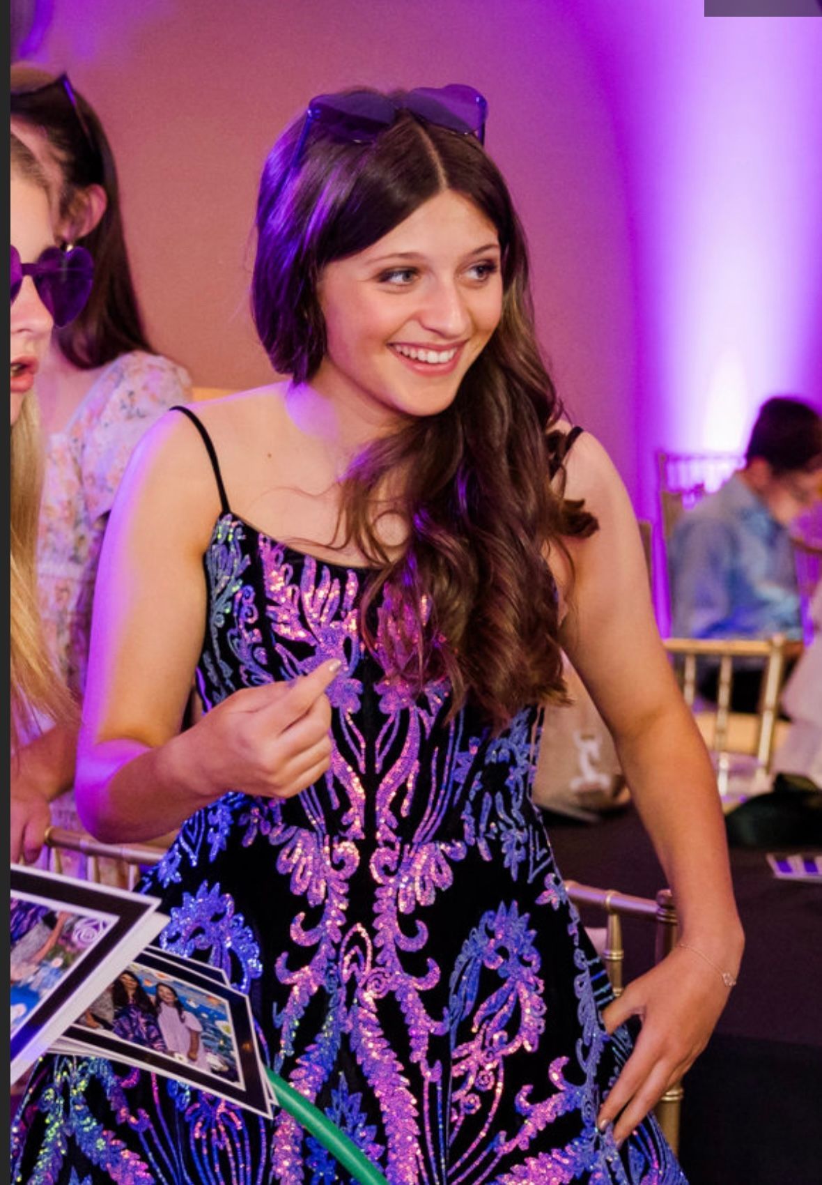 Young woman smiles, wearing a sequined black and purple dress. Party setting with others present.