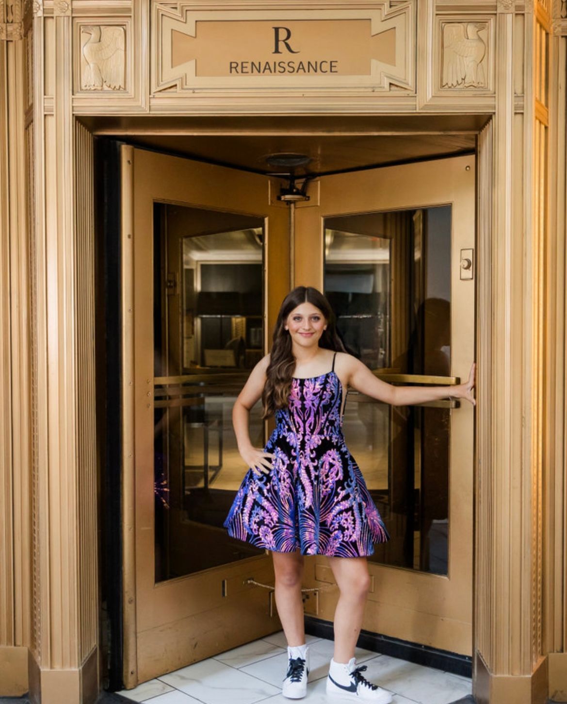 Girl in patterned dress stands in gold doorway of a Renaissance building, smiling.