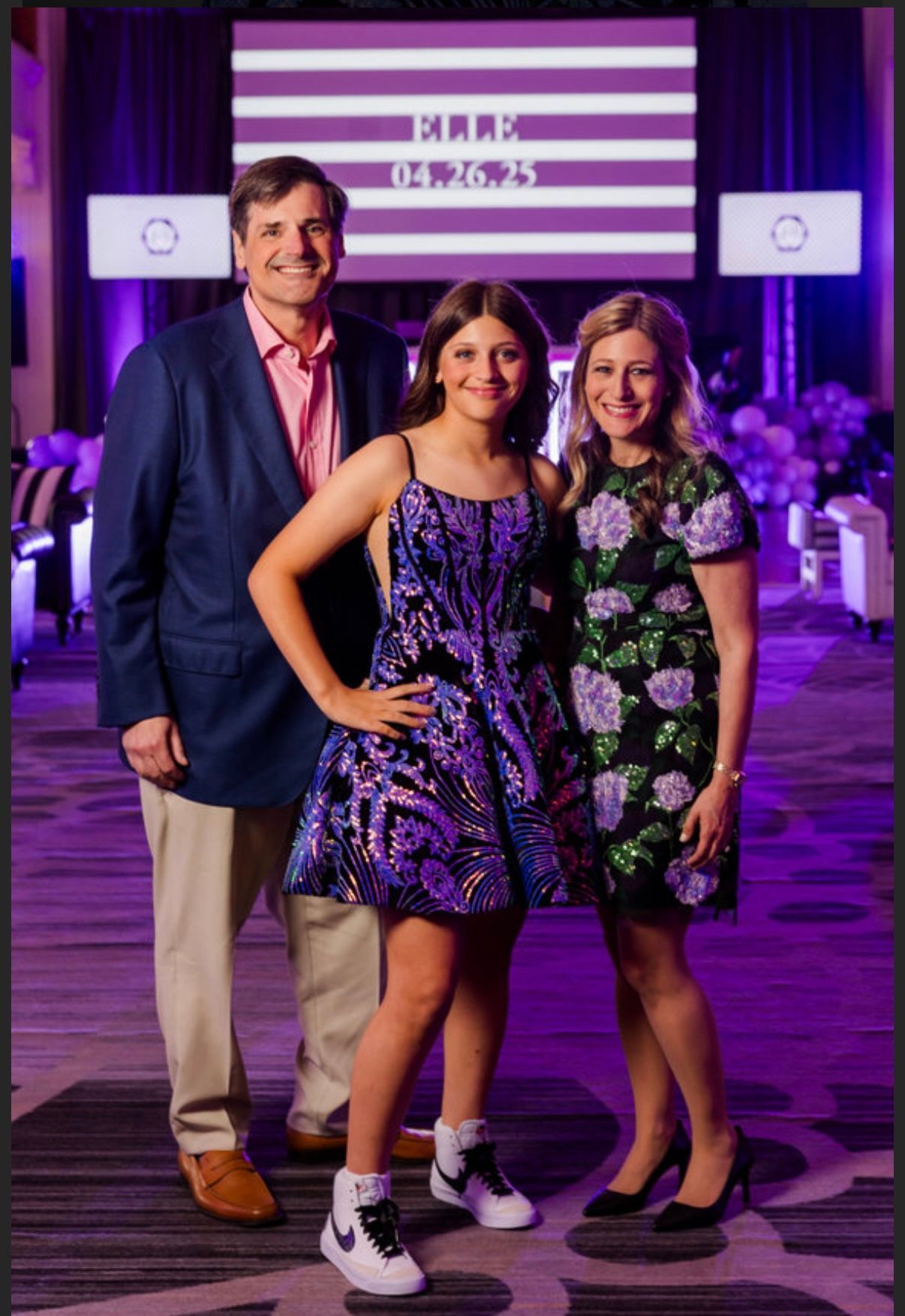 Family posing for photo at event; father, daughter in sequin dress and sneakers, and mother in floral dress. Purple setting.