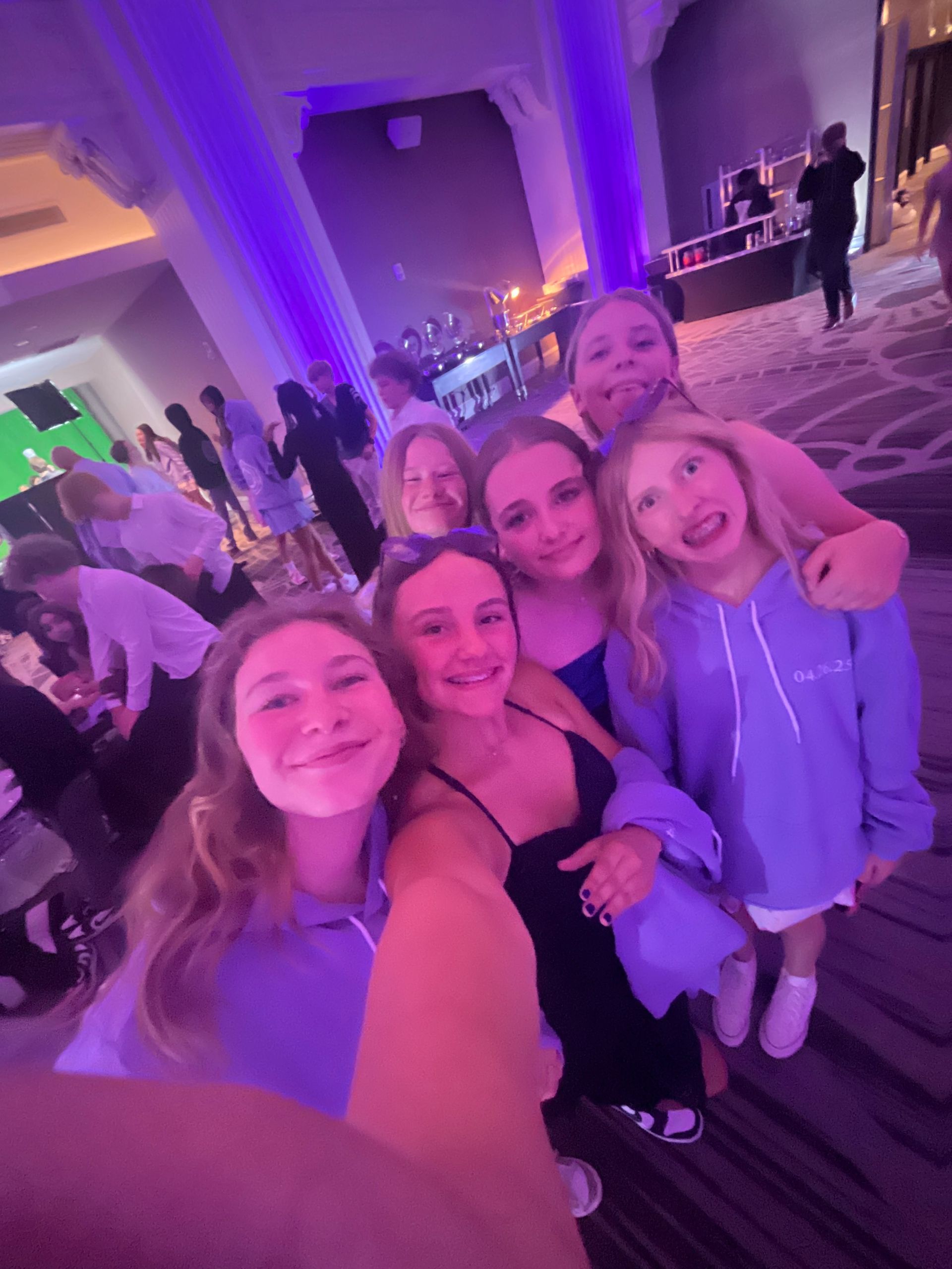 Group of girls smiling, hugging, taking a selfie at a party with purple lighting.