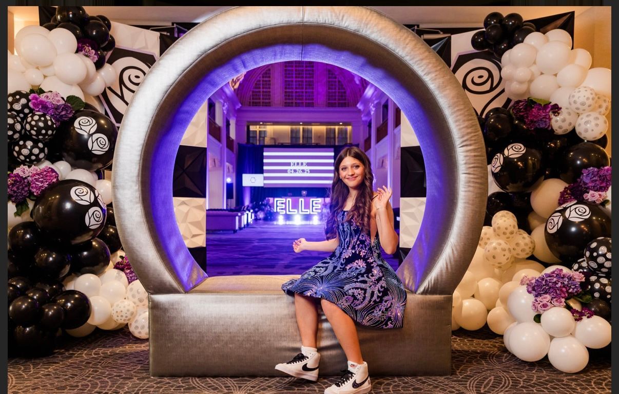 Girl in blue dress sits in silver inflatable arch, party with balloons.