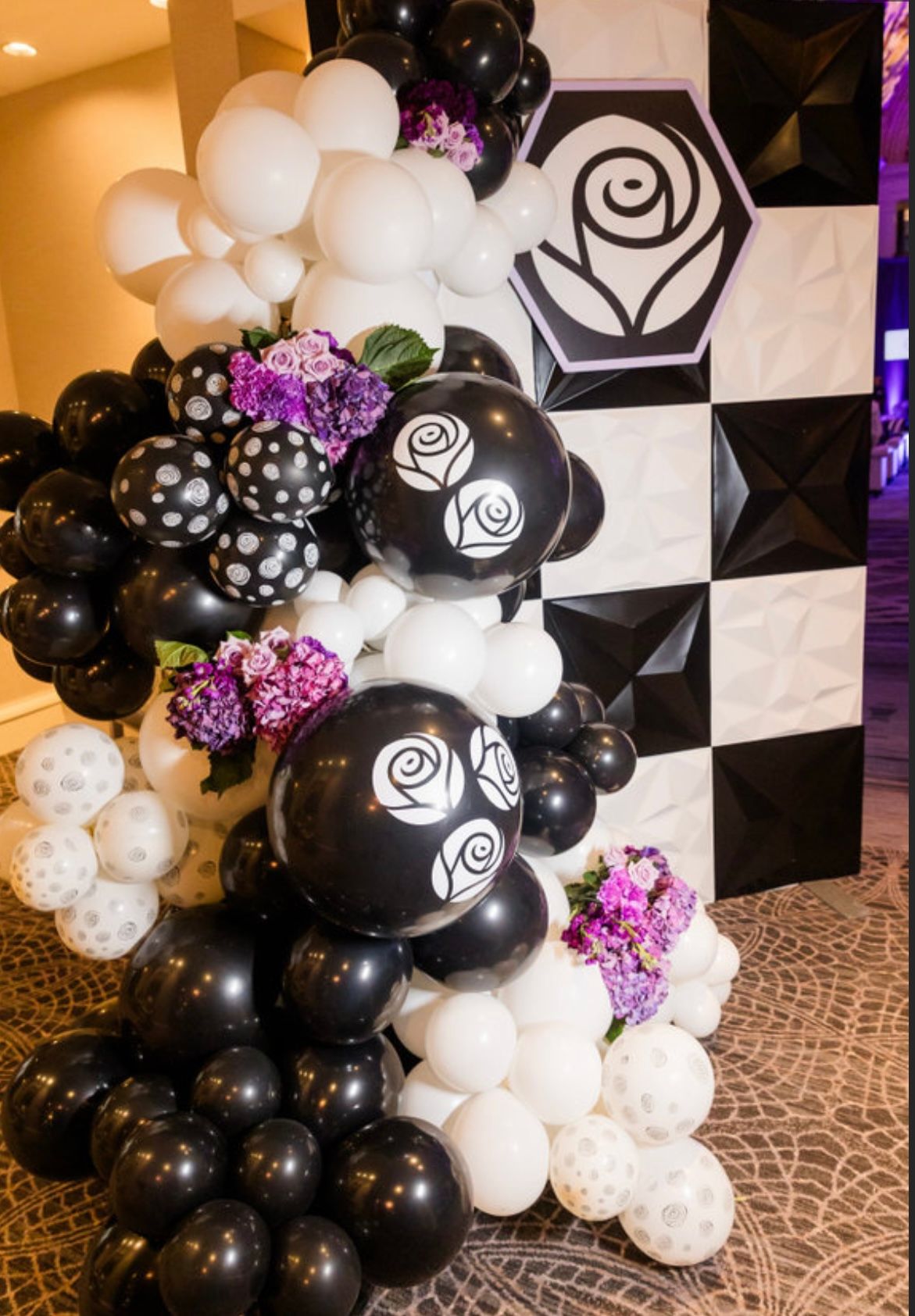 Black and white balloon arch with rose accents and purple flowers, beside a black and white wall with a rose logo.