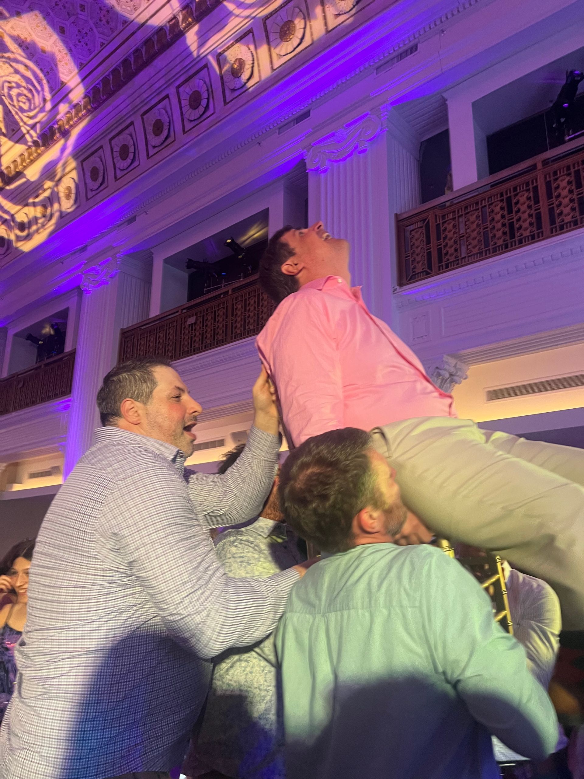 Men lift a man in a pink shirt. White columns and purple lighting in a grand ballroom setting.