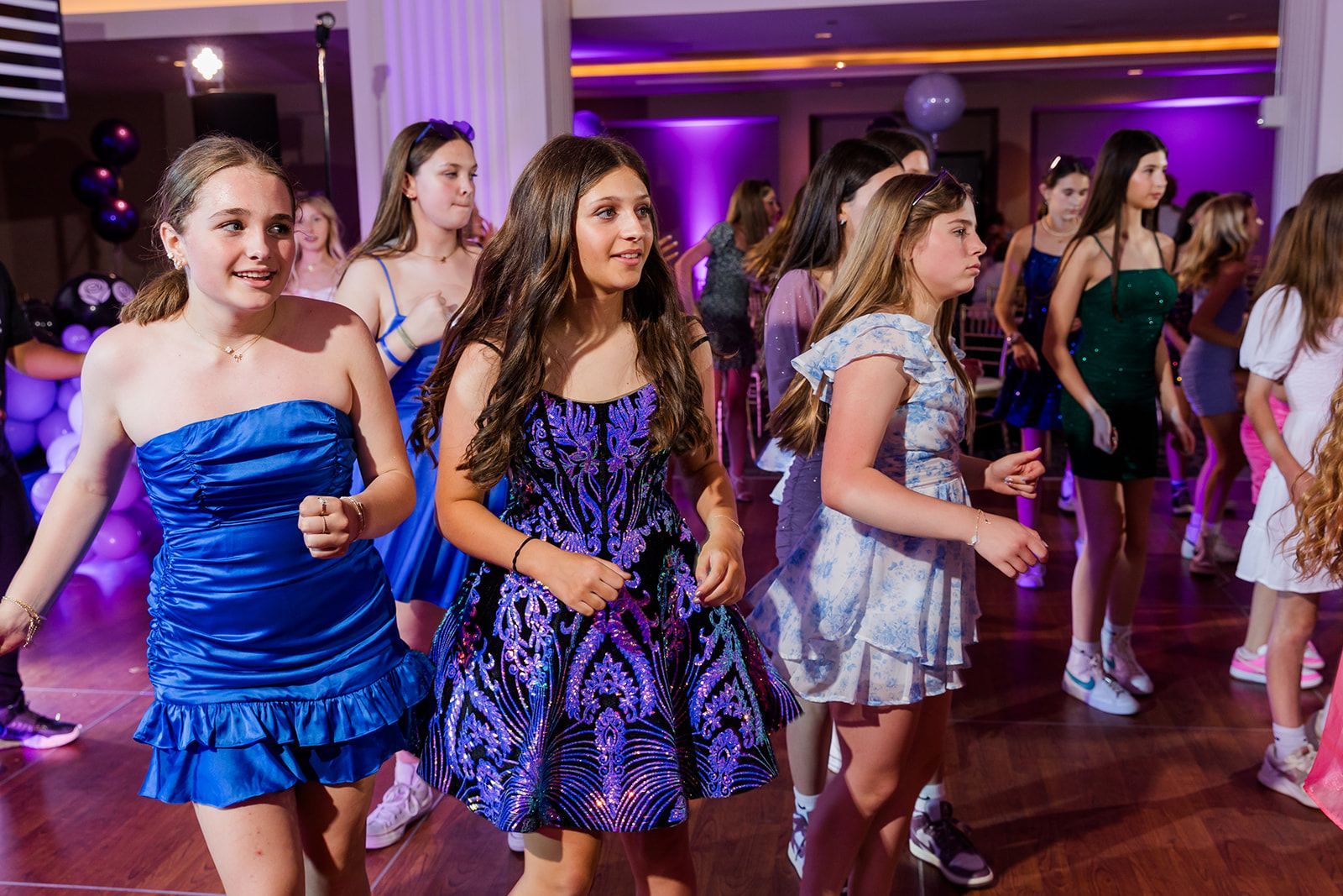 Girls dancing at a party, wearing dresses in blue, purple, and white. Bright, festive setting.