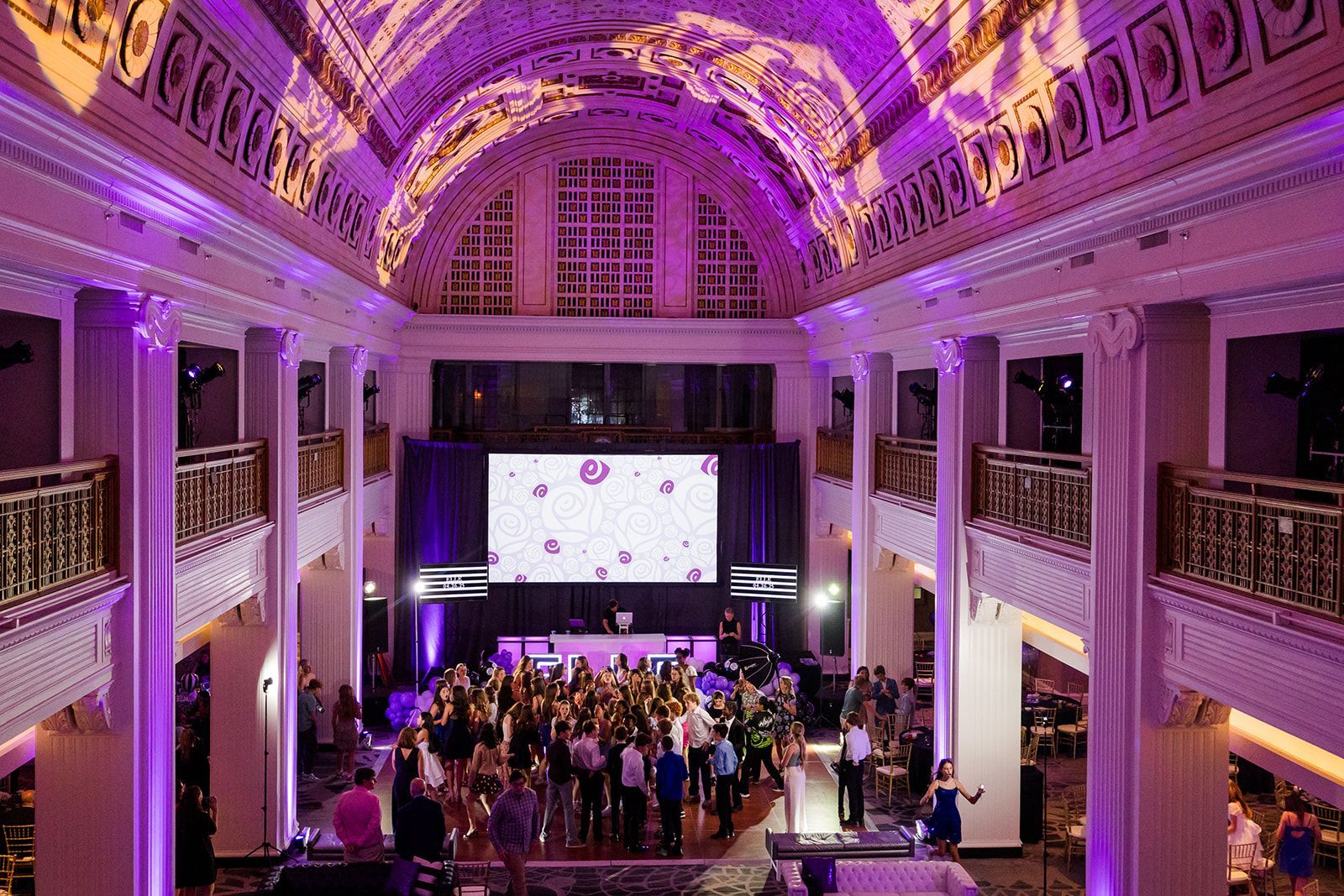 Large event space with a dance floor; purple and gold lighting; people dancing near a stage.