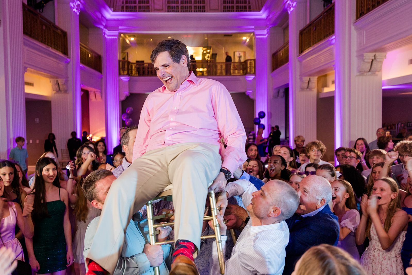 Man in pink shirt being lifted in a chair at a party with people cheering. Purple lighting, grand hall.