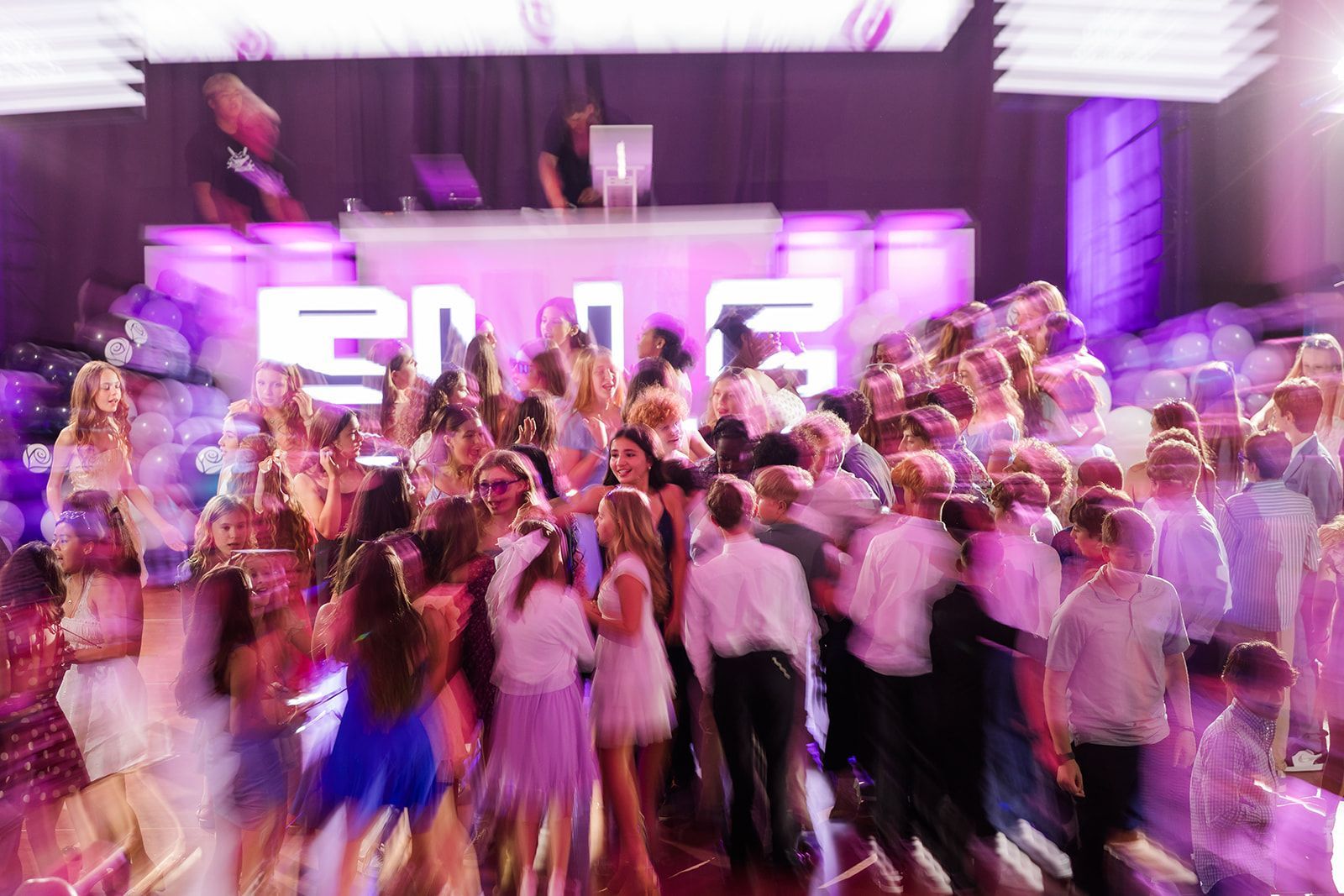 Blurred photo of a crowded dance floor with teens partying; purple and white lighting. DJ in the background.