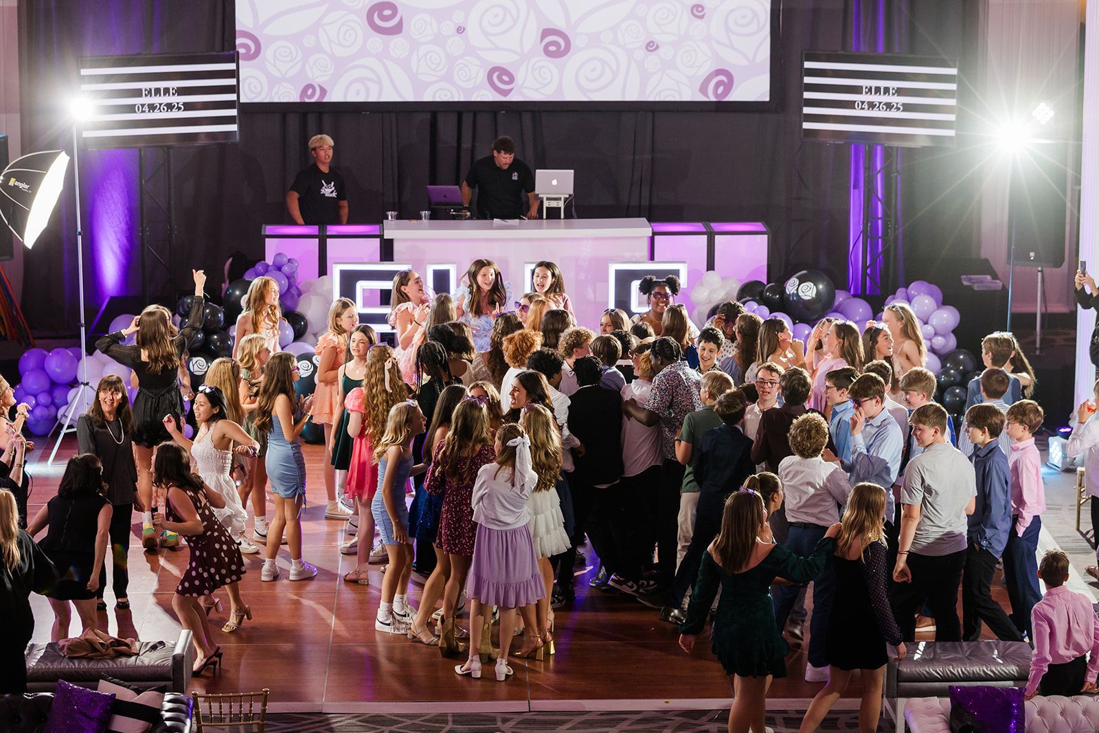 Teenagers dancing at a party with a DJ stage in front of a purple backdrop.