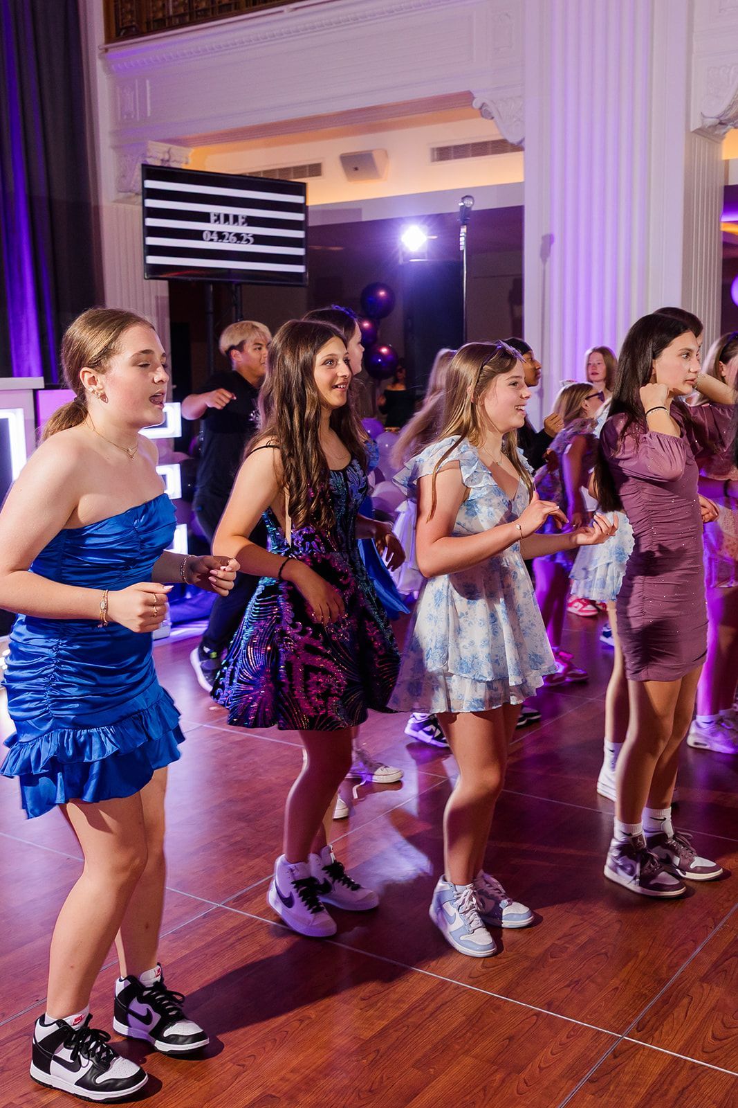 Girls dancing at a party, wearing dresses and sneakers. Lit room, DJ booth.