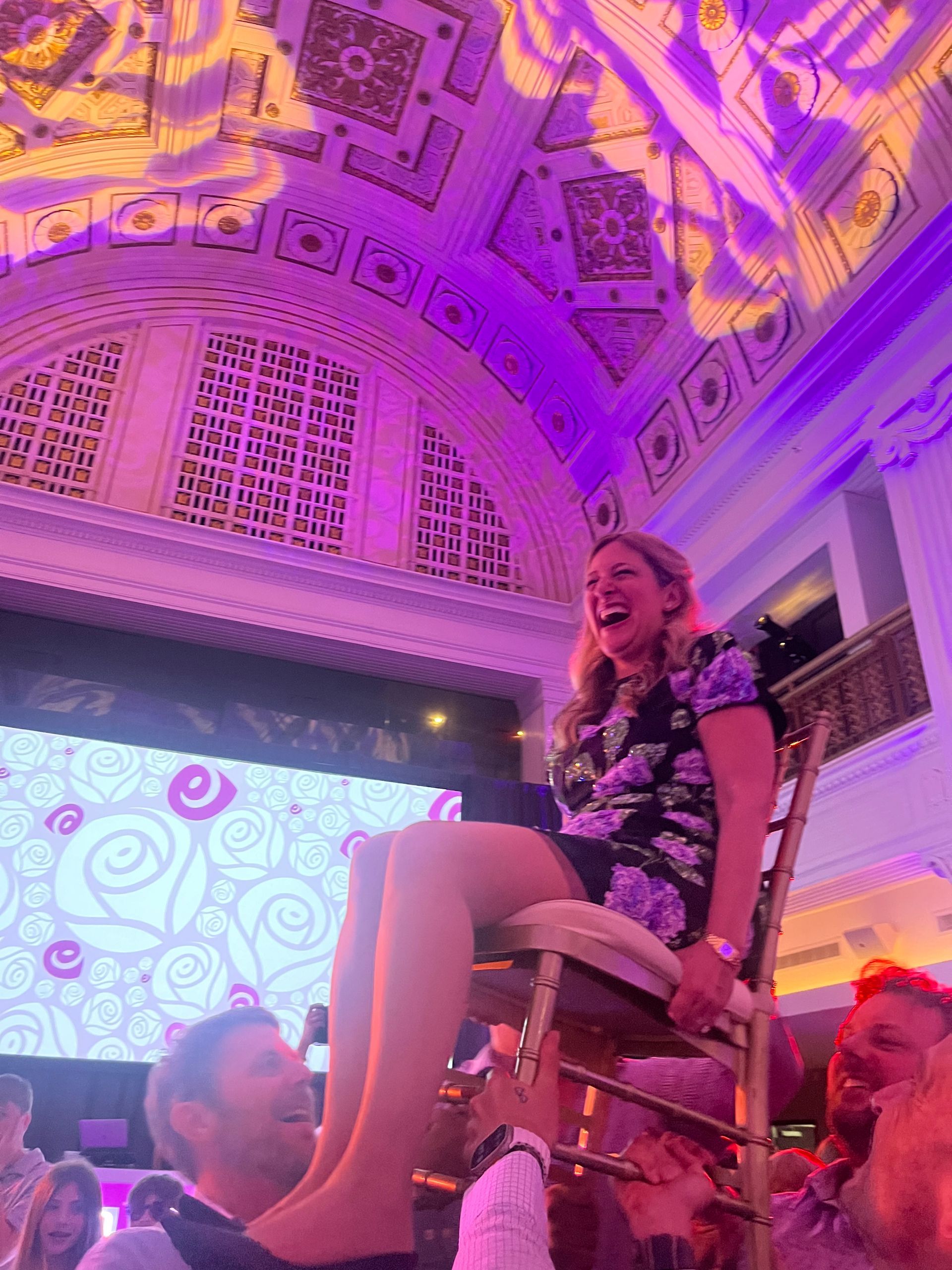 Woman on chair held high at celebration, laughing. Purple and gold lighting, ornate ceiling.