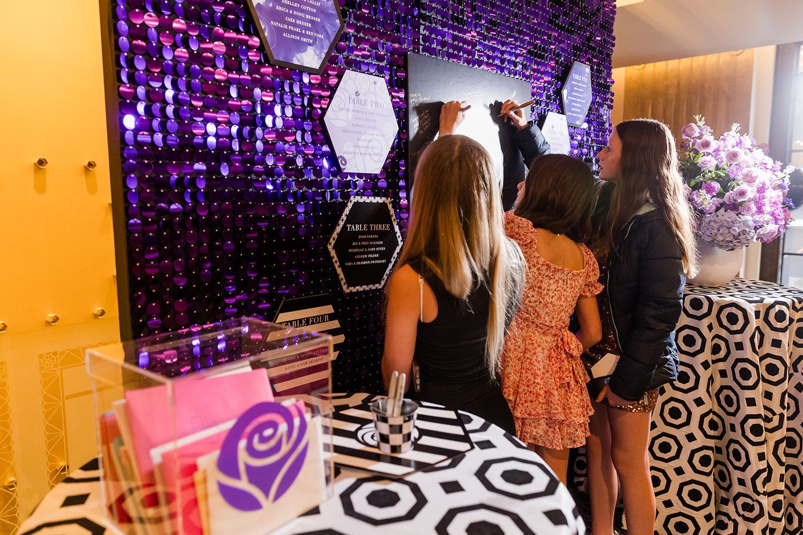 People signing a guest book with a purple sequined backdrop, a black and white patterned table, and a flower arrangement.