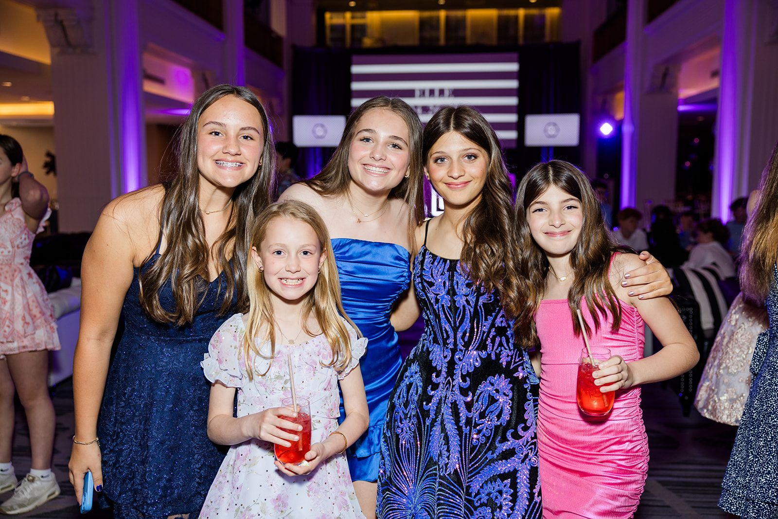 Five girls pose, smiling, at a party. They wear formal dresses in blue, pink, and purple, holding drinks.