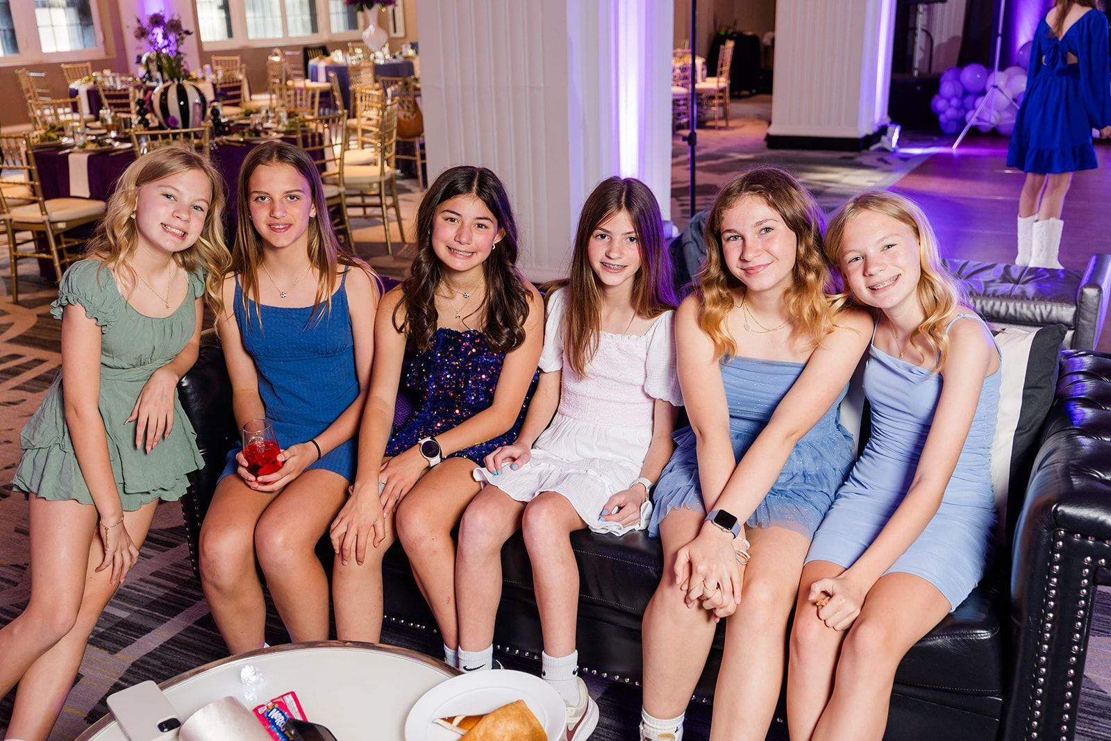 Group of six girls smiling, posing on a black couch at a party. They wear dresses, in a well-lit room.