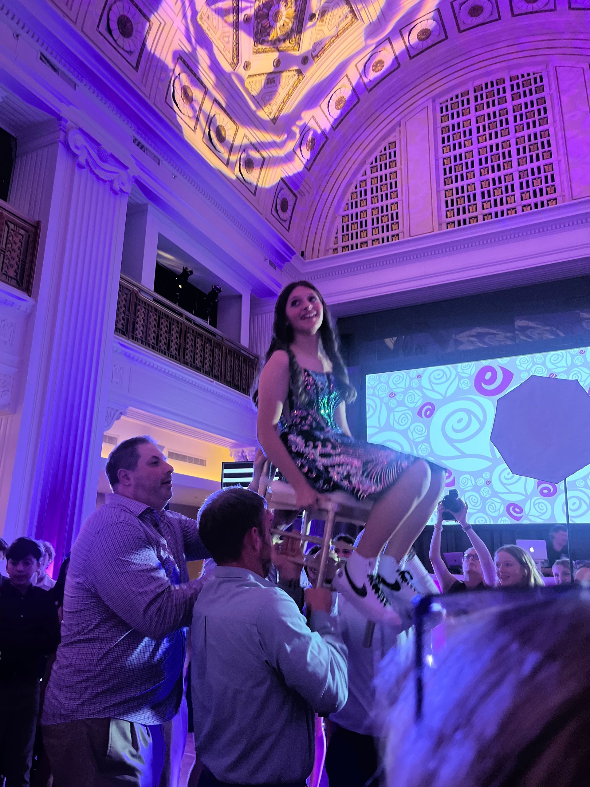 Teen girl on a chair held up by men in a purple-lit room, celebrating with a screen backdrop.
